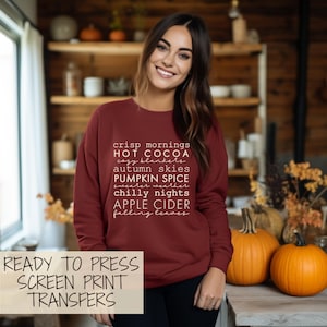 May include: A maroon sweatshirt with white text listing autumn-themed phrases like "crisp mornings" and "pumpkin spice." The words "Ready to Press Screen Print Transfers" are at the bottom. A woman smiles in the image, with pumpkins in the background.