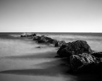 May include: A black and white photograph of a rocky breakwater extending into a misty, choppy sea.