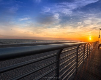 May include: A scenic boardwalk at sunset. The image features a metal railing in the foreground, leading the eye towards a wooden walkway and benches. The sky transitions from blue to orange and yellow, with the sun setting over the ocean.