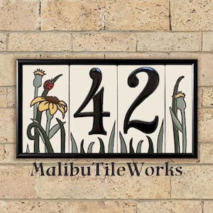 May include: A rectangular ceramic house number sign with the number "42" in glossy black. The sign features floral and grass designs, with a ladybug on a yellow flower. The text "MalibuTileWorks" is at the bottom. The sign is framed in black.