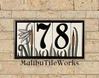 Puede incluir: Letrero rectangular con el número de casa 78 en negro sobre fondo blanco. El letrero presenta un diseño de libélula y espadañas. El letrero está enmarcado en negro y montado en una pared de ladrillo. El texto "MalibuTileWorks" está en la parte inferior.