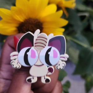May include: A cartoon cat-shaped enamel pin with large eyes and a light beige body. The pin has black and burgundy accents on the ears and eyes. The pin is held in front of a yellow sunflower.