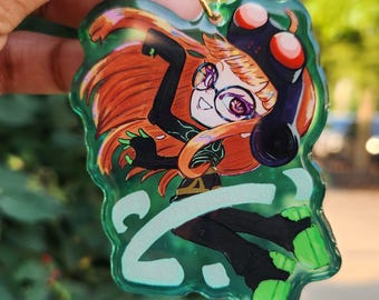 May include: A green acrylic keychain featuring a character with orange hair, glasses, and a black outfit. The keychain has a gold star-shaped clasp. The character is set against a green background with a stylized design.