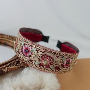 May include: A burgundy headband with gold and floral embroidery. The headband has a detailed pattern of flowers and sequins, creating an ornate and decorative accessory. The headband is approximately 2 cm wide.