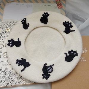 May include: A white beret with black embroidered cats. The beret has a round shape and is made of a soft, fuzzy material.
