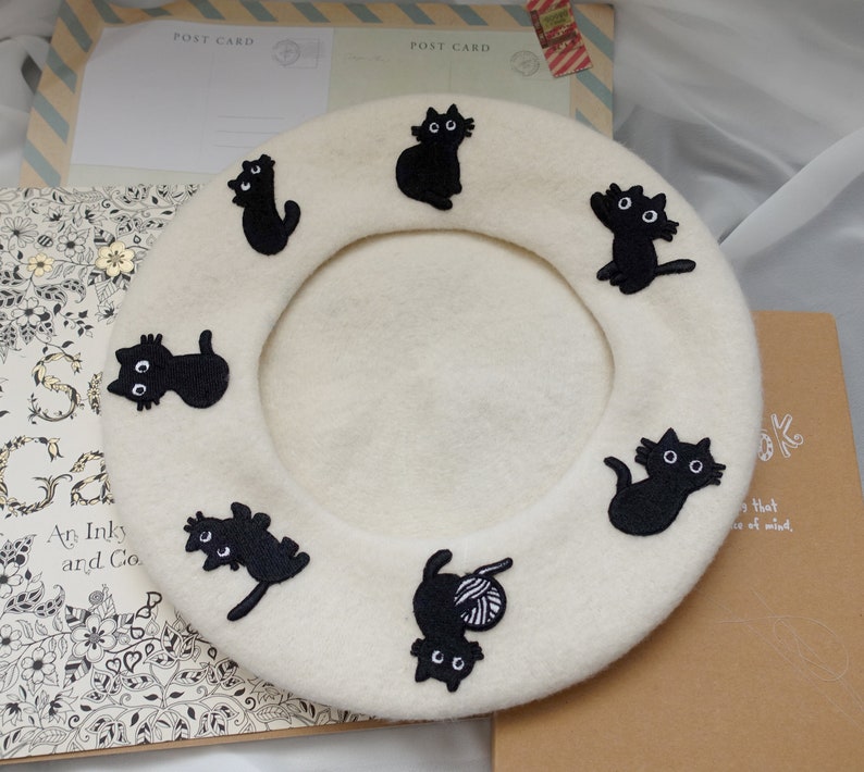 May include: A white beret with black embroidered cats. The beret has a round shape and is made of a soft, fuzzy material.