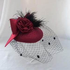 Op de afbeelding: Een bordeauxrode fascinator met een zwarte gestippelde sluier. De hoed heeft een grote stoffen roos en zwarte veren. De hoofdbedekking heeft een kleine rand en een decoratieve strik. Dit elegante accessoire is geschikt voor speciale gelegenheden.