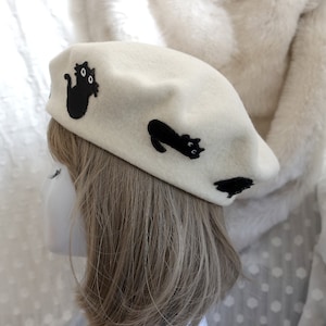 May include: A white felt beret with black embroidered cats. The beret has a classic shape and is decorated with three black cats, each with a unique pose and expression.