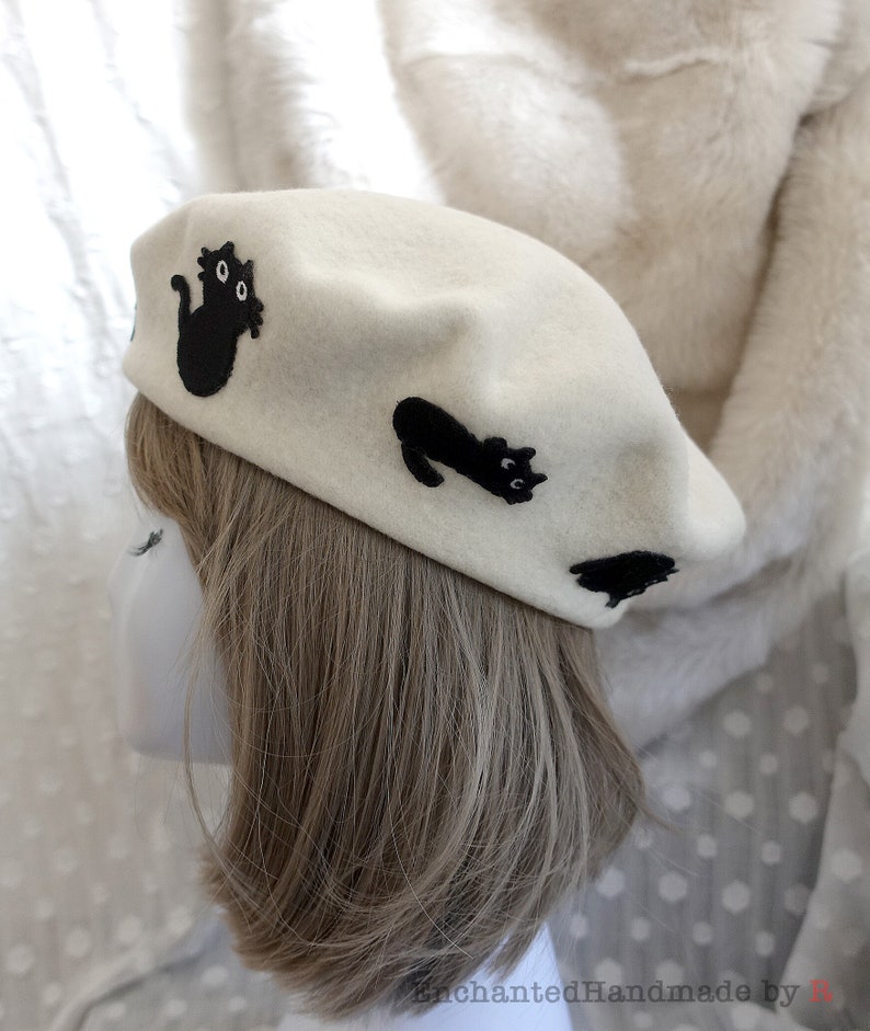 May include: A white felt beret with black embroidered cats. The beret has a classic shape and is decorated with three black cats, each with a unique pose and expression.