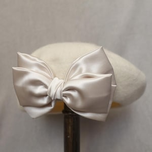May include: A cream-colored beret with a large, satin bow in a matching shade. The bow is prominently displayed on the front of the hat, adding a touch of elegance. The beret is made of a soft, felt-like material.