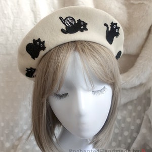May include: A white beret with black embroidered cats. One cat is holding a ball of yarn.