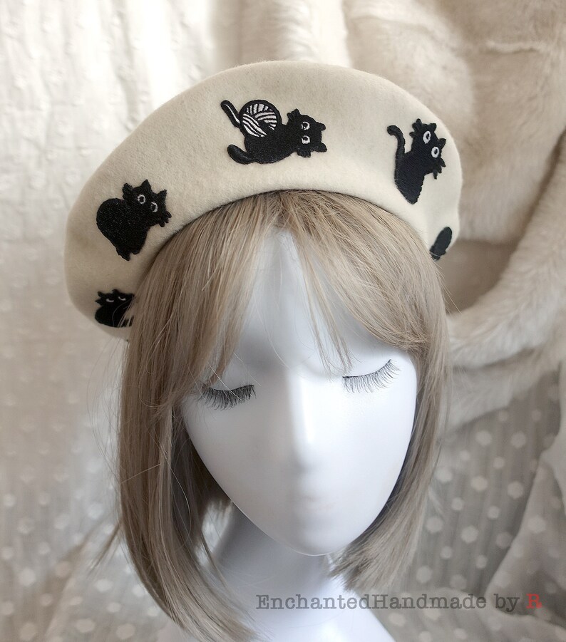 May include: A white beret with black embroidered cats. One cat is holding a ball of yarn.