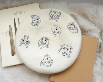 May include: A white beret with black embroidered dog illustrations. The beret is made of a soft, fuzzy material.