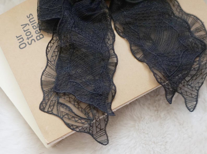 May include: Close-up of a dark navy blue lace ribbon with a scalloped edge, resting on a book. The book has the text "Our Story Begins" printed on the cover. The lace has a delicate, intricate pattern.