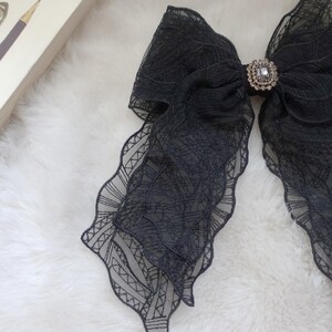May include: A large, black lace bow with a decorative rhinestone center. The bow has a layered design with scalloped edges. The bow is on a white, fluffy surface.