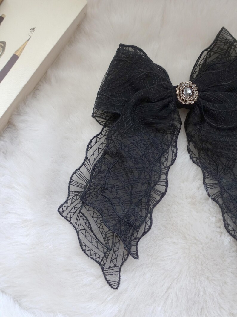 May include: A large, black lace bow with a decorative rhinestone center. The bow has a layered design with scalloped edges. The bow is on a white, fluffy surface.