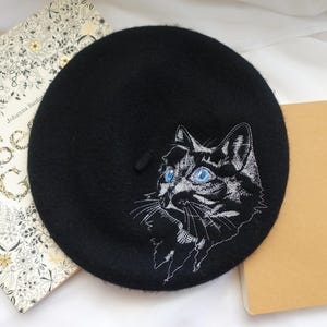 以下が含まれることがあります： 青い目をした白い刺繍の猫のデザインが施された黒いベレー帽