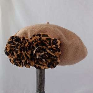 May include: A tan felt beret featuring two large, layered fabric flowers with a brown and black leopard print pattern attached to the side. The beret has a soft, rounded shape and a smooth, uniform texture across the crown.