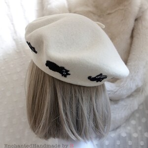 May include: A white beret with black embroidered cats. The beret is made of felt and has a classic shape.