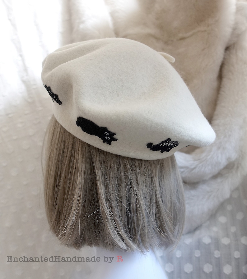May include: A white beret with black embroidered cats. The beret is made of felt and has a classic shape.