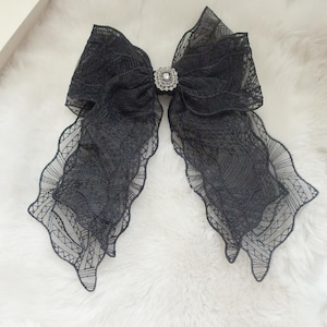 May include: A large black lace bow with a decorative rhinestone center. The bow has a layered design with scalloped edges, creating a delicate and elegant appearance. The bow is set against a fluffy white background.