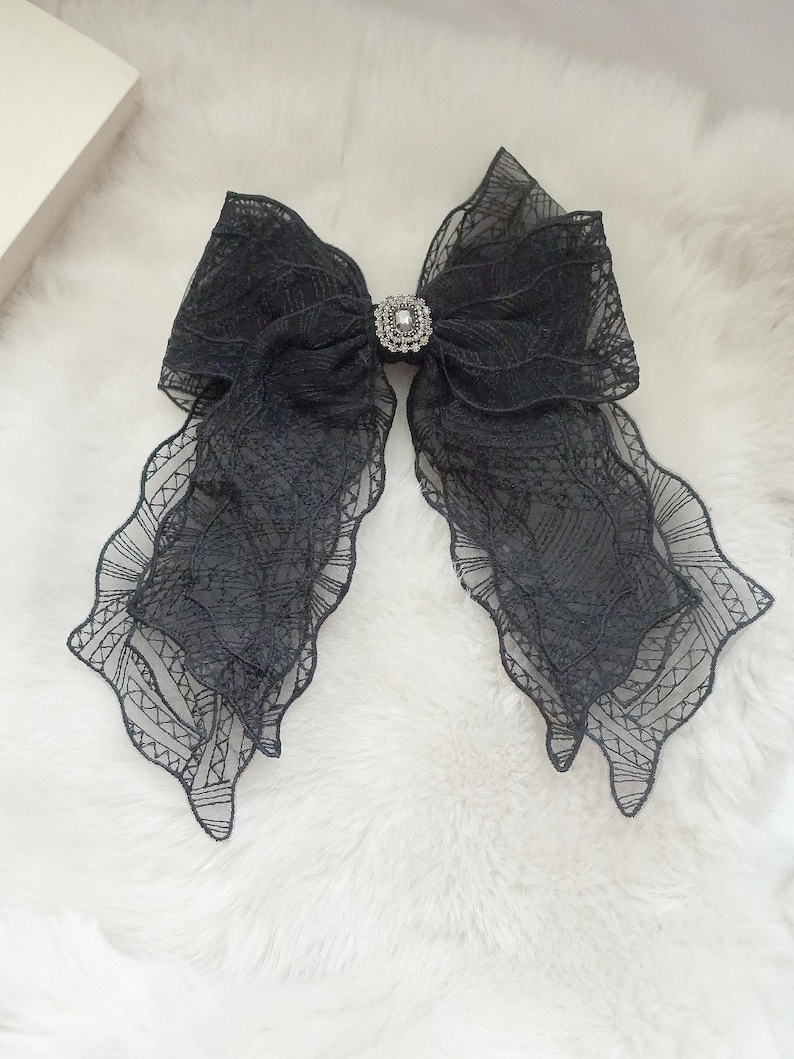 May include: A large black lace bow with a decorative rhinestone center. The bow has a layered design with scalloped edges, creating a delicate and elegant appearance. The bow is set against a fluffy white background.