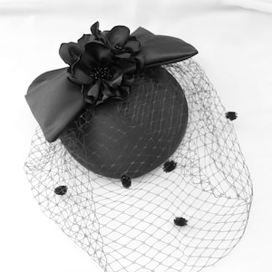 Puede incluir: Un sombrero fascinador negro con un adorno floral y un lazo. El sombrero presenta un velo delicado con pequeños adornos decorativos. Un accesorio formal para ocasiones especiales.