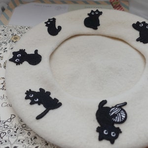 May include: A white beret with black embroidered cats. The cats are in various poses, including sitting, standing, and playing with yarn.