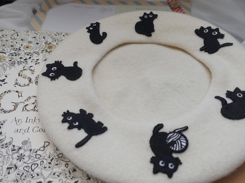 May include: A white beret with black embroidered cats. The cats are in various poses, including sitting, standing, and playing with yarn.