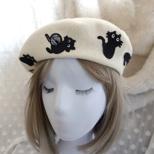May include: A white beret with black embroidered cats. One cat is holding a ball of yarn.