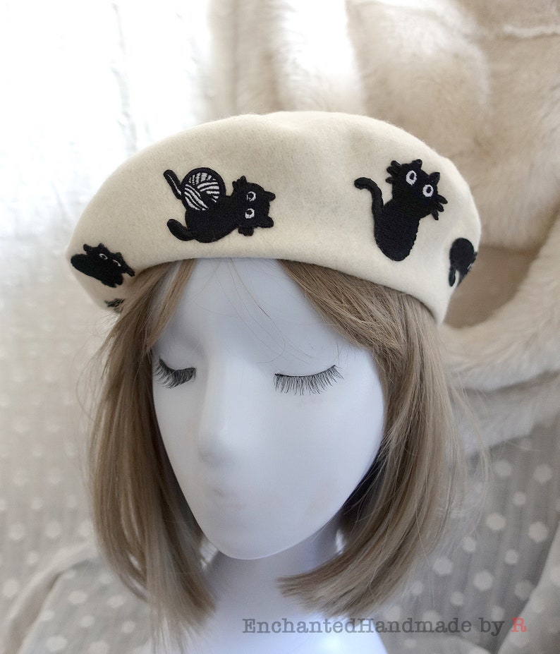 May include: A white beret with black embroidered cats. One cat is holding a ball of yarn.