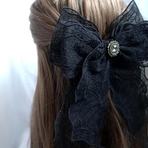 May include: A large black lace bow hair accessory with a decorative rhinestone center. The bow is attached to the hair, with the back of the head and brown hair visible.