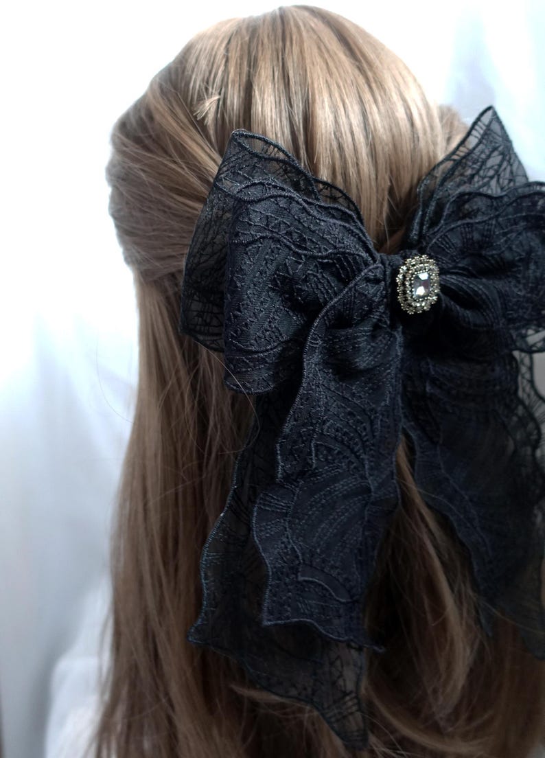 May include: A large black lace bow hair accessory with a decorative rhinestone center. The bow is attached to the hair, with the back of the head and brown hair visible.