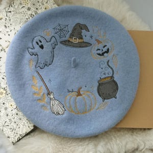 May include: A light blue beret with embroidered Halloween-themed designs. The beret features a ghost, witch's hat, pumpkin, cauldron, spiderweb, and broom. The embroidery is in black, gold, and orange. The beret is made of a soft, felt-like material.