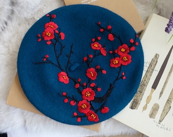 May include: A teal blue beret with black embroidered branches and red flowers. The flowers have yellow centers.