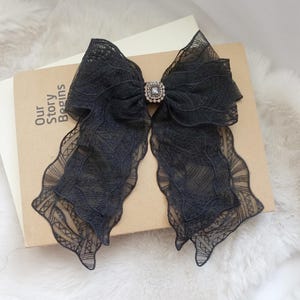 May include: A black lace bow with a rhinestone center, resting on a book. The bow has long, flowing tails with a scalloped edge. The book has the words "Our Story Begins" printed on the cover.