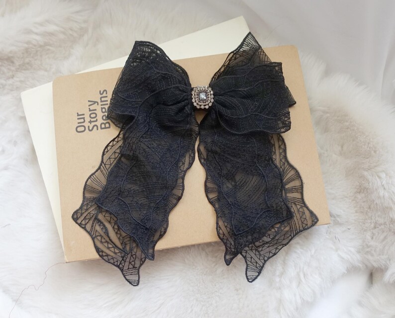 May include: A black lace bow with a rhinestone center, resting on a book. The bow has long, flowing tails with a scalloped edge. The book has the words "Our Story Begins" printed on the cover.
