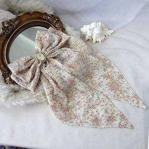 Könnte beinhalten: Eine große Haarschleife aus cremefarbenem Stoff mit einem zarten rosa-grünen Blumenmuster und goldfarbenen Stickranken. In der Mitte der Schleife befindet sich eine ovale Brosche mit einem weißen, kameeartigen Blumenmotiv und kleinen Perlen.