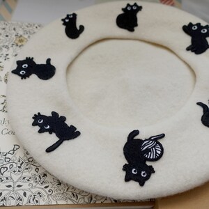 May include: A white beret with black embroidered cats. The cats are in various poses, including sitting, standing, and curled up.