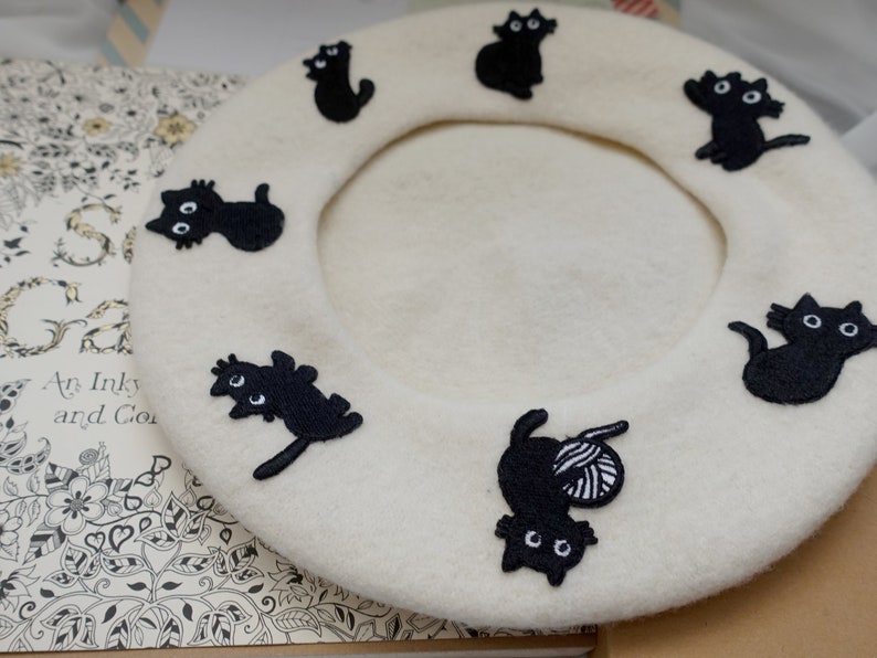 May include: A white beret with black embroidered cats. The cats are in various poses, including sitting, standing, and curled up.