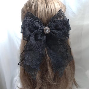 May include: A large black lace bow hair clip with a sparkling rhinestone center. The bow is attached to a barrette and is worn in the hair. The hair is light brown.