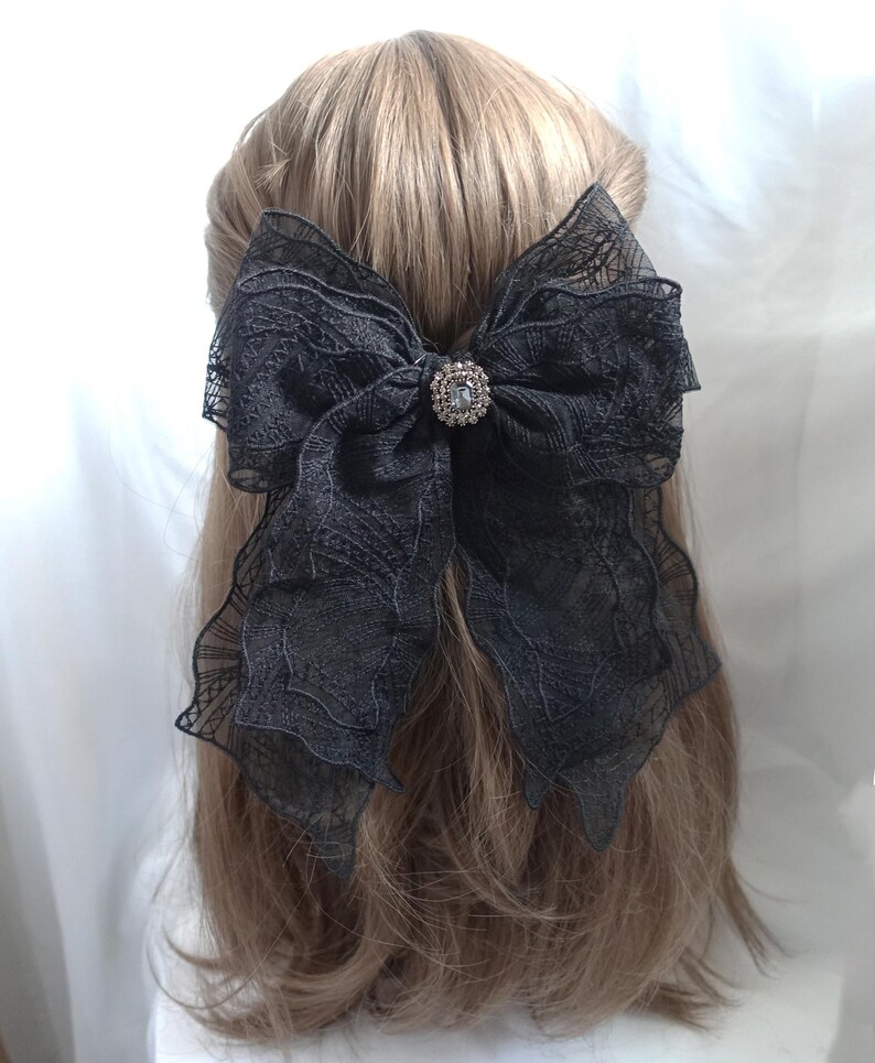 May include: A large black lace bow hair clip with a sparkling rhinestone center. The bow is attached to a barrette and is worn in the hair. The hair is light brown.