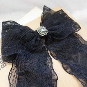 May include: A black lace bow with a decorative rhinestone center. The bow is made of delicate, sheer fabric with intricate patterns. The bow is a hair accessory.