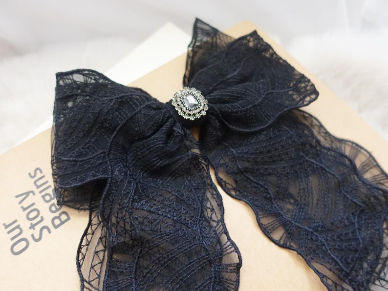 May include: A black lace bow with a decorative rhinestone center. The bow is made of delicate, sheer fabric with intricate patterns. The bow is a hair accessory.
