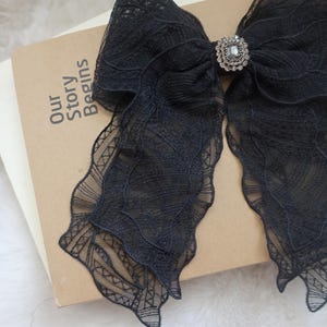 May include: A large black lace bow with a decorative rhinestone center. The bow is resting on a book with the words "Our Story Begins" printed on the cover. The lace has a delicate, patterned design.