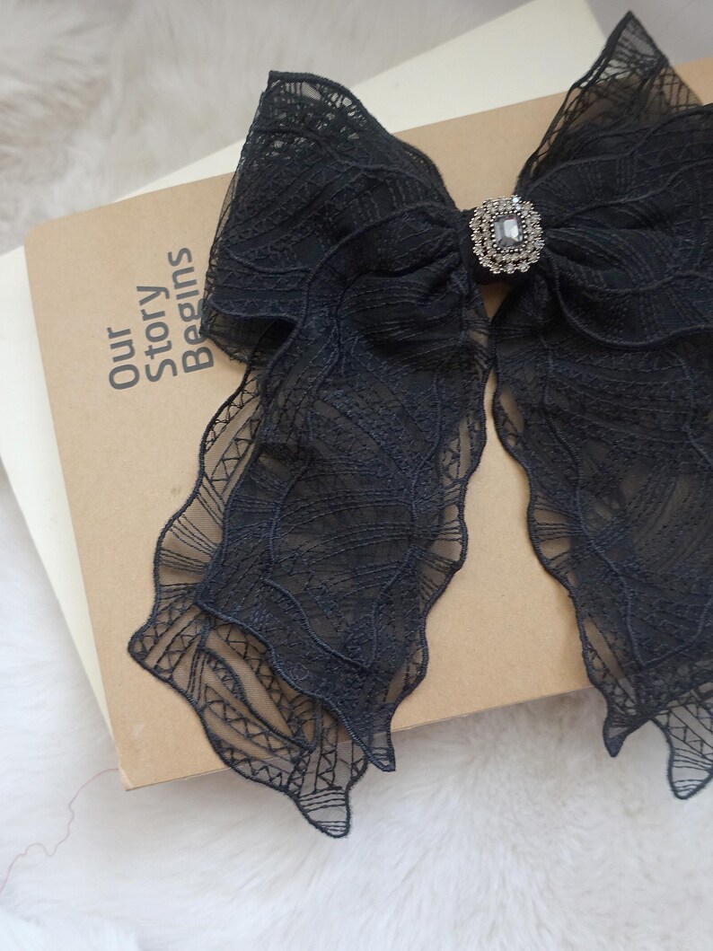 May include: A large black lace bow with a decorative rhinestone center. The bow is resting on a book with the words "Our Story Begins" printed on the cover. The lace has a delicate, patterned design.