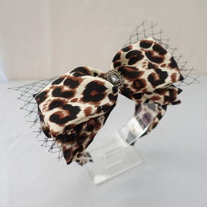Könnte beinhalten: Ein Haarband mit Leopardenmuster, mit einer großen Schleife und einem schwarzen Netzschleier. Die Schleife ist cremefarben, braun und schwarz. Ein dekorativer Schmuck befindet sich in der Mitte der Schleife. Das Haarband steht auf einem transparenten Displayständer.