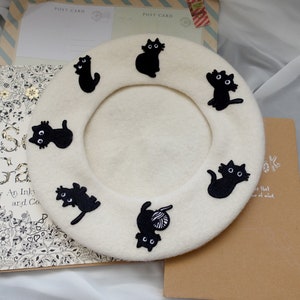May include: A white beret with black embroidered cats around the brim. The cats are in various poses, including sitting, standing, and playing with yarn.
