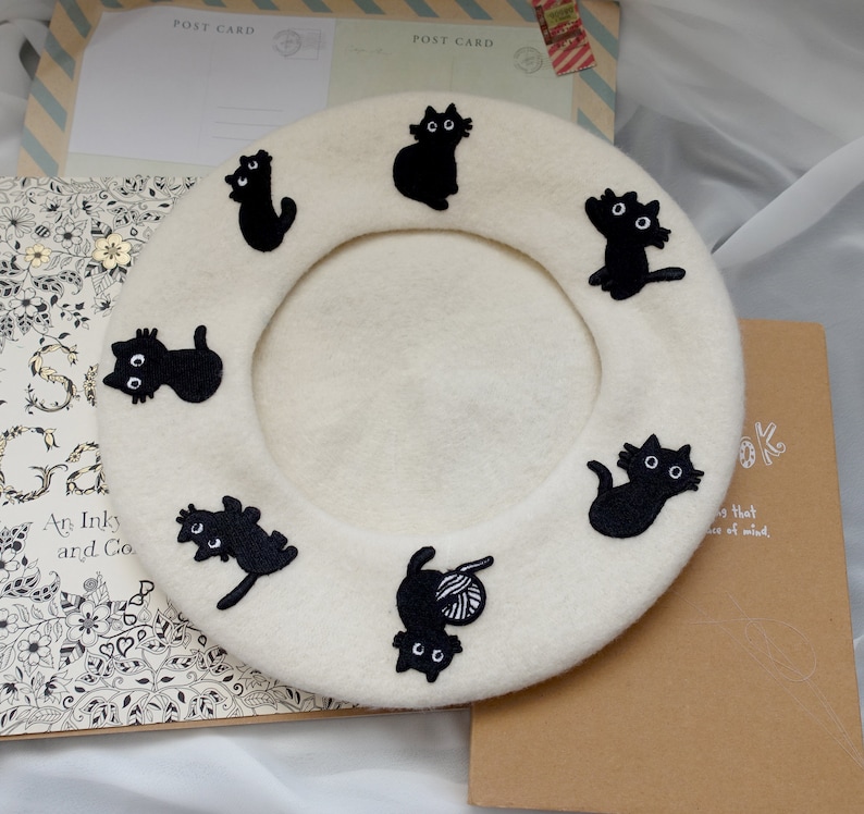 May include: A white beret with black embroidered cats around the brim. The cats are in various poses, including sitting, standing, and playing with yarn.