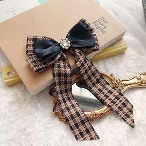 May include: A hair bow with a dark blue satin centre and a jewel. The bow is set on a brown and black houndstooth fabric. Long, matching fabric streamers extend from the bow. The bow is a stylish accessory.